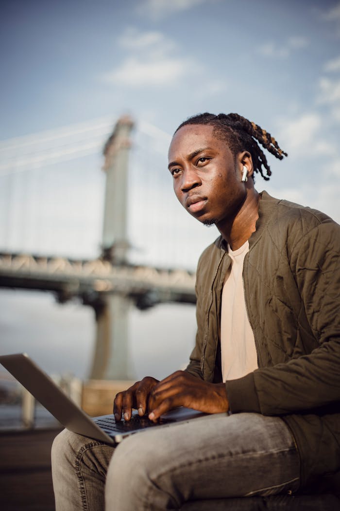 Serious African American male employee in casual clothes working outdoors and using laptop while sitting in park at city centre